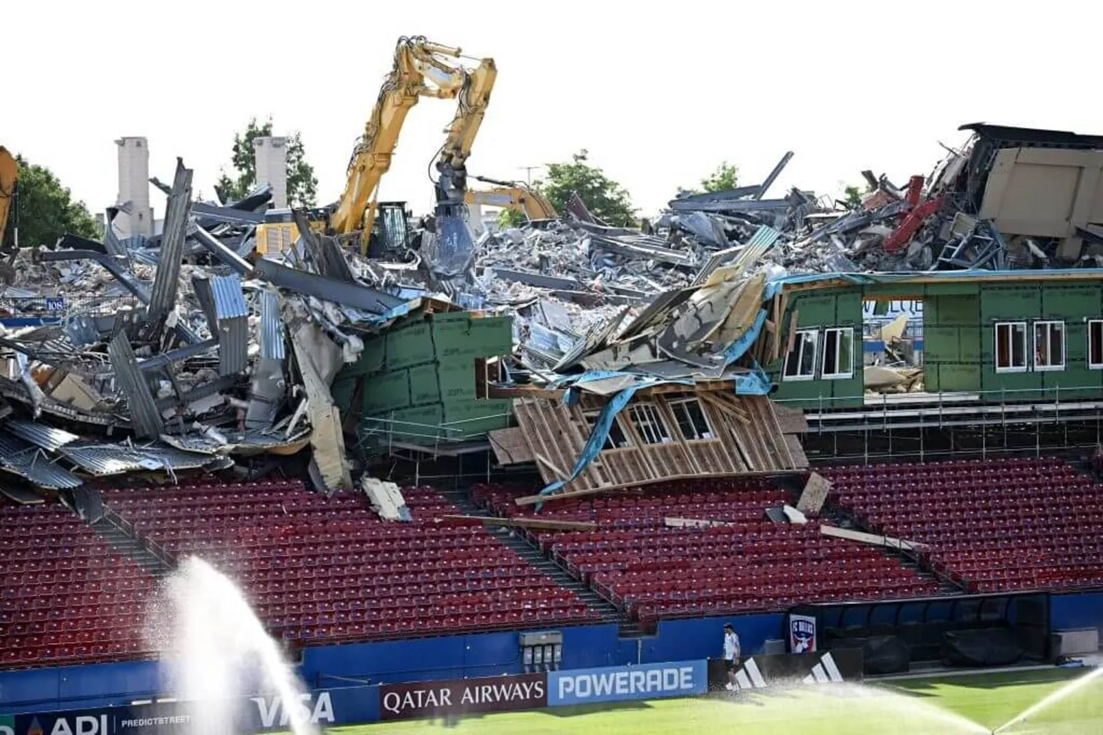 A large digger towers over the crumbling grandstand of FC Dallas' stadium, while a sprinkler waters the grass below
