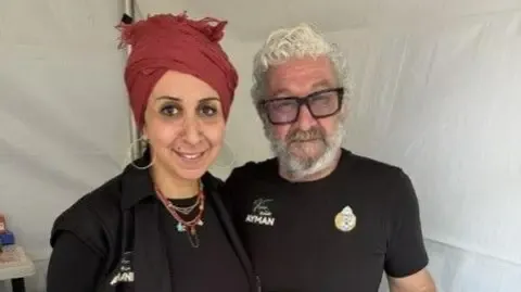 A man and woman stand in a temporary pop-up food stall, smiling, she has her hair wrapped in a red scarf while he has thick black glasses and silver hair and beard.
