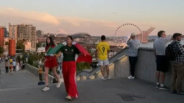Football fans in different colour kits take in the view of the Seattle skyline from a high vantage point with a giant ferris wheel in the background.